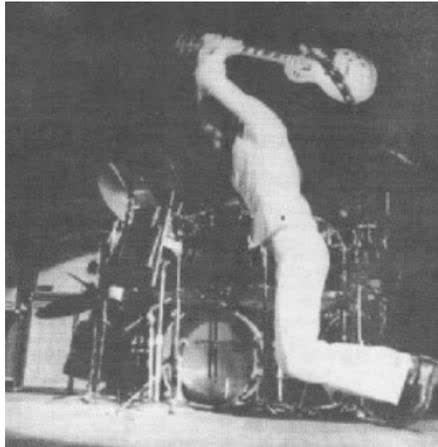 A black and white photograph of a man in a white suit, possibly a musician, performing on stage. He is captured in a dynamic pose, leaning forward with his arms raised, suggesting movement or dancing. The background is dark and indistinct, typical of a stage setting.